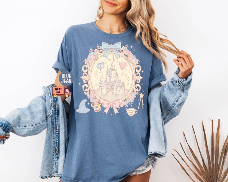 K&ouml;nnte beinhalten: Ein hellblaues T-Shirt mit einer pastellrosa und blauen Illustration von Schloss Aschenputtel, Mickey-Maus-Ohren und einer Teetasse. Die Illustration ist von einem Blumengirlanden und einer Schleife umgeben.