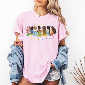 Puede incluir: Camiseta rosa con un gr&aacute;fico de siete princesas de dibujos animados con vestidos coloridos y un fondo de estrellas. Las princesas miran hacia la derecha y est&aacute;n en fila.