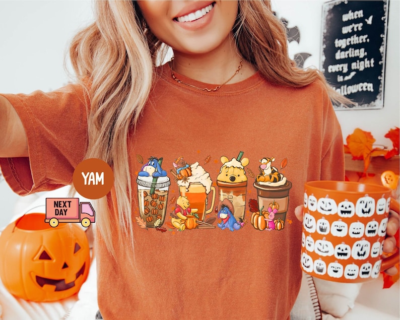 May include: Burnt orange t-shirt featuring a line of cartoon characters with fall-themed drinks. The characters include Winnie the Pooh, Eeyore, and Tigger. A pumpkin-themed mug is held in the hand. The shirt has a round neck.