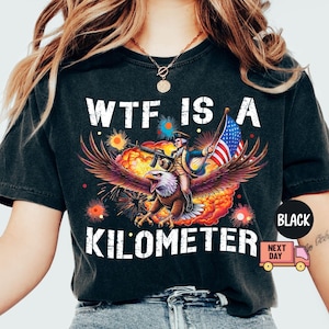May include: Black graphic t-shirt with the text "WTF IS A KILOMETER". The design features an eagle carrying a person with a gun and an American flag, set against a fiery background.