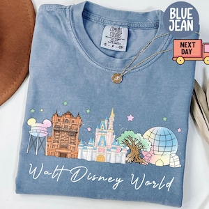 May include: Blue jean-colored t-shirt with a graphic of Walt Disney World landmarks. The design includes a water tower, Tower of Terror, Cinderella Castle, Spaceship Earth, and the text "Walt Disney World". The shirt has a crew neck and short sleeves.