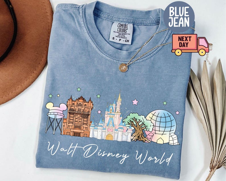 May include: Blue jean-colored t-shirt with a graphic of Walt Disney World landmarks. The design includes a water tower, Tower of Terror, Cinderella Castle, Spaceship Earth, and the text "Walt Disney World". The shirt has a crew neck and short sleeves.