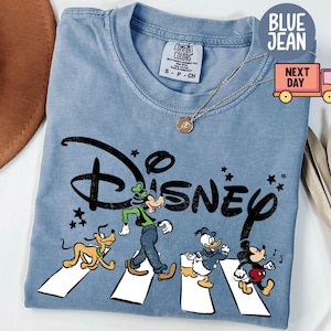 K&ouml;nnte beinhalten: Hellblaues T-Shirt mit dem Wort "Disney" in Schwarz und den Zeichentrickfiguren Goofy, Pluto, Donald Duck und Mickey Mouse, die einen wei&szlig;en Zebrastreifen &uuml;berqueren. Das Shirt hat einen Rundhalsausschnitt und kurze &Auml;rmel.