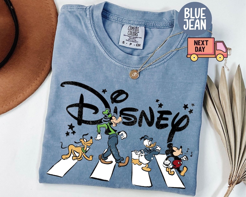 K&ouml;nnte beinhalten: Hellblaues T-Shirt mit dem Wort "Disney" in Schwarz und den Zeichentrickfiguren Goofy, Pluto, Donald Duck und Mickey Mouse, die einen wei&szlig;en Zebrastreifen &uuml;berqueren. Das Shirt hat einen Rundhalsausschnitt und kurze &Auml;rmel.