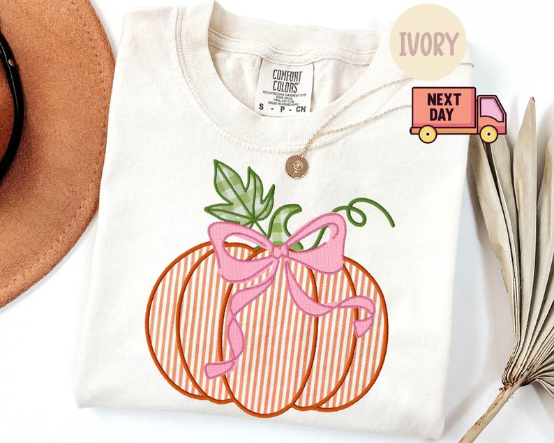 May include: Cream-coloured Comfort Colors t-shirt with an embroidered pumpkin design. The pumpkin has orange and white stripes, a pink bow, and green leaves. A gold necklace and a brown hat are also visible.