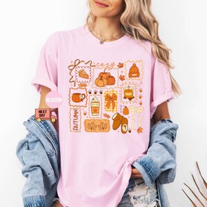May include: A light pink t-shirt featuring a collection of autumn-themed illustrations. The design includes pumpkins, a coffee cup, a candle, and the word "cozy" in a decorative font. The word "Autumn" is also present. The shirt is a relaxed fit.