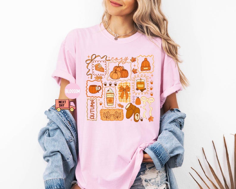 May include: A light pink t-shirt featuring a collection of autumn-themed illustrations. The design includes pumpkins, a coffee cup, a candle, and the word "cozy" in a decorative font. The word "Autumn" is also present. The shirt is a relaxed fit.