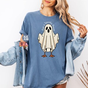 May include: Blue t-shirt with a graphic of a ghost-goose. The ghost is white with black eyes and a beak, standing on orange feet. The text "Just a Spooky Goose" is arched above the ghost. The shirt has rolled-up sleeves.