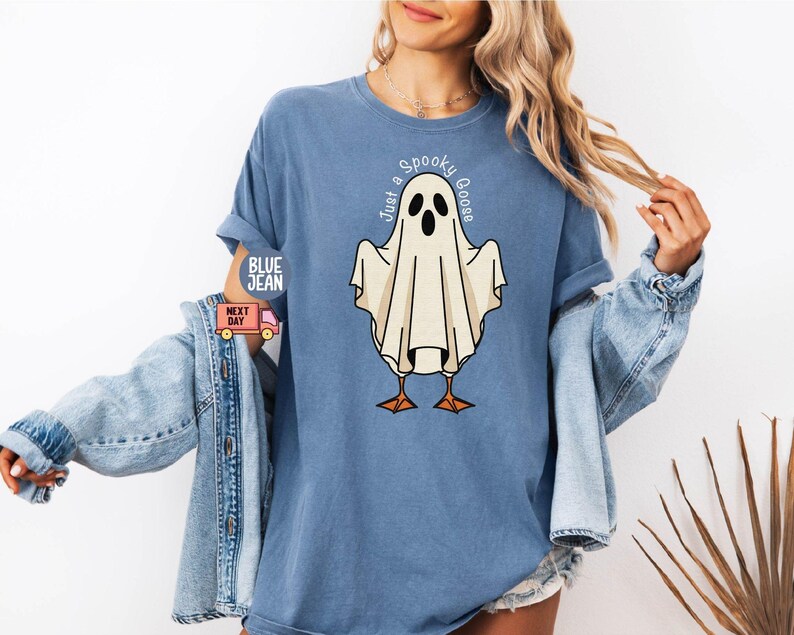 May include: Blue t-shirt with a graphic of a ghost-goose. The ghost is white with black eyes and a beak, standing on orange feet. The text "Just a Spooky Goose" is arched above the ghost. The shirt has rolled-up sleeves.