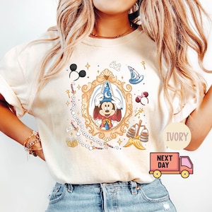 Könnte beinhalten: Elfenbeinfarbenes T-Shirt mit einer gerahmten Illustration von Mickey Mouse mit Zaubererhut. Das Design enthält Disney-Elemente wie Micky-Maus-Ohren, einen Zauberstab und einen Zaubererhut. Das Shirt hat eine lockere Passform.