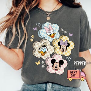 May include: Dark gray t-shirt featuring a floral design with Disney characters Daisy Duck, Donald Duck, Minnie Mouse, and Mickey Mouse. The flowers have checkered patterns in pastel colors. Butterflies and small flowers are also included in the design.