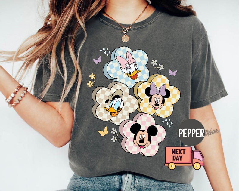 May include: Dark gray t-shirt featuring a floral design with Disney characters Daisy Duck, Donald Duck, Minnie Mouse, and Mickey Mouse. The flowers have checkered patterns in pastel colors. Butterflies and small flowers are also included in the design.