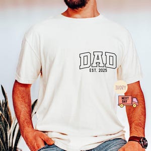 May include: Ivory-colored t-shirt with the word "DAD" in a collegiate font, and the text "EST. 2025" below it. The shirt is short-sleeved and has a relaxed fit. A small graphic of a delivery truck is visible on the lower right.