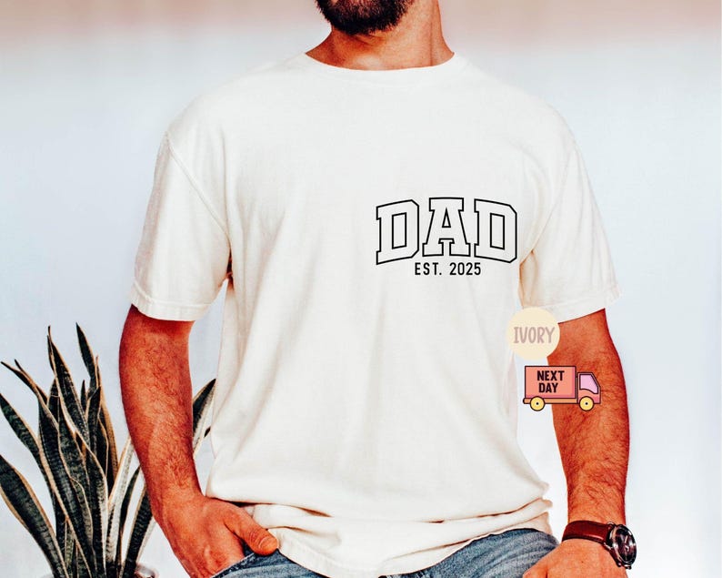 May include: Ivory-colored t-shirt with the word "DAD" in a collegiate font, and the text "EST. 2025" below it. The shirt is short-sleeved and has a relaxed fit. A small graphic of a delivery truck is visible on the lower right.