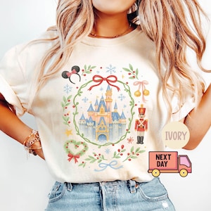 Puede incluir: Camiseta color marfil con una ilustración de un castillo de cuento de hadas, rodeada de elementos festivos como una corona, lazos y un cascanueces. El diseño incluye orejas de ratón y copos de nieve, evocando un tema navideño.