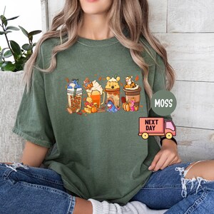 May include: Green t-shirt featuring a colorful graphic of Winnie the Pooh characters as fall-themed beverages. The design includes Eeyore, Pooh, Piglet, Tigger, and other characters. The shirt has a relaxed fit and is made of a soft material.