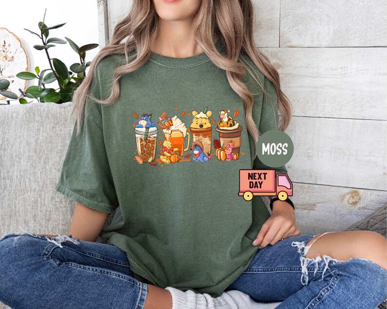 May include: Green t-shirt featuring a colorful graphic of Winnie the Pooh characters as fall-themed beverages. The design includes Eeyore, Pooh, Piglet, Tigger, and other characters. The shirt has a relaxed fit and is made of a soft material.