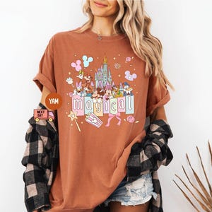 May include: A rust-colored t-shirt featuring a colorful graphic of Disney characters, a castle, and the word "magical". The design includes Mickey and Minnie Mouse, Donald Duck, and other characters. The shirt has rolled-up sleeves.