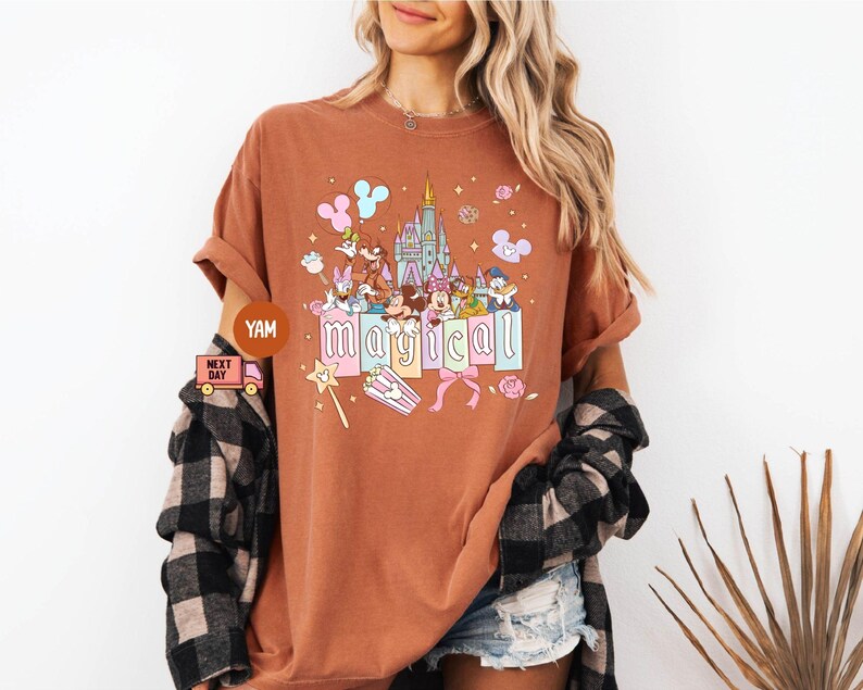 May include: A rust-colored t-shirt featuring a colorful graphic of Disney characters, a castle, and the word "magical". The design includes Mickey and Minnie Mouse, Donald Duck, and other characters. The shirt has rolled-up sleeves.