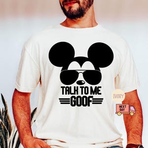May include: An ivory short-sleeved t-shirt featuring a black graphic of a mouse silhouette wearing sunglasses. Below the graphic, the text reads TALK TO ME GOOF in bold black capital letters, flanked by two horizontal lines on each side of the word GOOF.