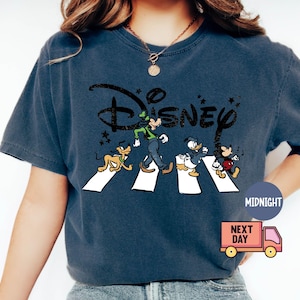 K&ouml;nnte beinhalten: Marineblaues T-Shirt mit einem Disney-Design mit Goofy, Pluto, Donald Duck und Mickey Mouse, die eine Stra&szlig;e &uuml;berqueren. Das Disney-Logo befindet sich &uuml;ber den Figuren. Das T-Shirt hat einen Vintage-Look.