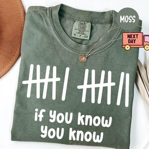 May include: A moss green t-shirt with white text reading "if you know you know" and a tally mark design. The shirt has a crew neck and short sleeves. A gold necklace is draped across the shirt.