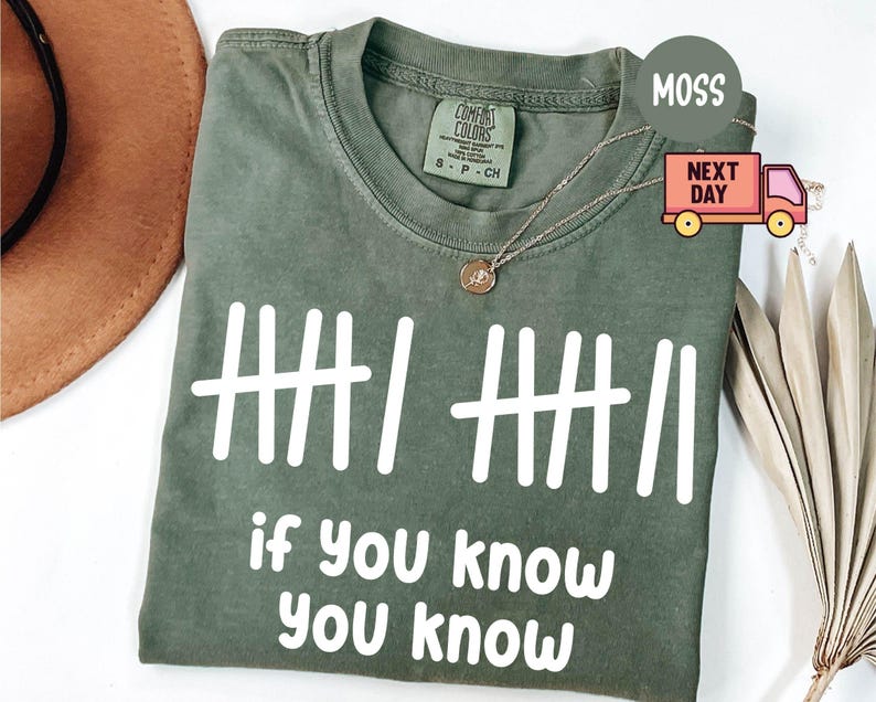May include: A moss green t-shirt with white text reading "if you know you know" and a tally mark design. The shirt has a crew neck and short sleeves. A gold necklace is draped across the shirt.