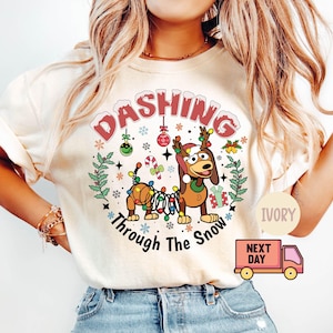 May include: A white t-shirt with a Christmas-themed design featuring a cartoon dog wearing a Santa hat and tangled in Christmas lights. The text "Dashing Through The Snow" is printed below the dog. The shirt is available in ivory.