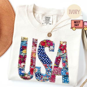 May include: Ivory-colored Comfort Colors t-shirt with the word "USA" in a patchwork design. The letters are filled with floral and star-spangled patterns in red, blue, and pink. A gold necklace and a brown hat are also visible.