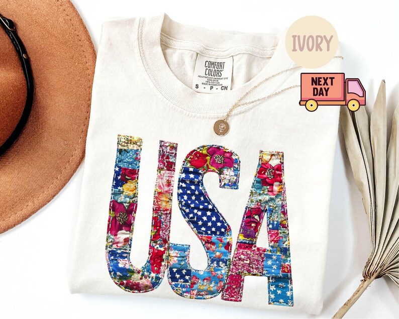 May include: Ivory-colored Comfort Colors t-shirt with the word "USA" in a patchwork design. The letters are filled with floral and star-spangled patterns in red, blue, and pink. A gold necklace and a brown hat are also visible.