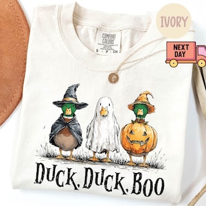 May include: Ivory-colored t-shirt featuring three ducks dressed in Halloween costumes. One duck is a witch, another a ghost, and the third is a jack-o'-lantern. The text "Duck. Duck. Boo" is printed below the ducks.