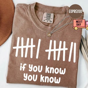 May include: Espresso-colored t-shirt with white graphic text and tally marks. The text reads "if you know, you know." The shirt has a crew neck and short sleeves. A silver necklace with a pendant is visible.