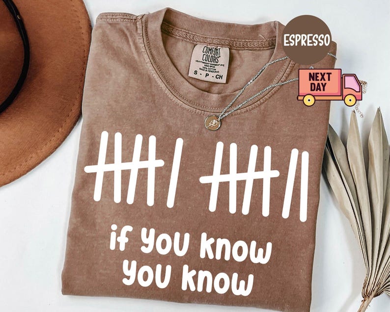May include: Espresso-colored t-shirt with white graphic text and tally marks. The text reads "if you know, you know." The shirt has a crew neck and short sleeves. A silver necklace with a pendant is visible.
