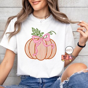 May include: White t-shirt with a pumpkin design, pink bow, and green leaves. The pumpkin has orange vertical stripes. The word "WHITE" is printed on a white circle. The design is a seasonal theme.