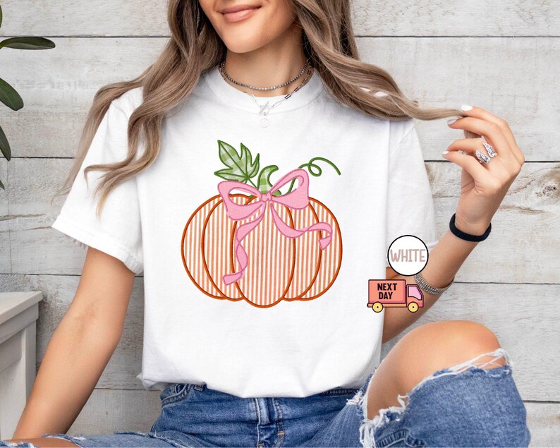 May include: White t-shirt with a pumpkin design, pink bow, and green leaves. The pumpkin has orange vertical stripes. The word "WHITE" is printed on a white circle. The design is a seasonal theme.