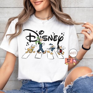 K&ouml;nnte beinhalten: Wei&szlig;es T-Shirt mit einem Disney-Motiv. Das Design zeigt Goofy, Pluto, Donald Duck und Mickey Mouse auf einem Zebrastreifen, mit dem Disney-Logo dar&uuml;ber. Das T-Shirt hat einen klassischen Rundhalsausschnitt.