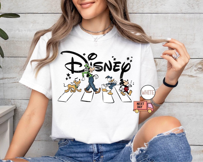 K&ouml;nnte beinhalten: Wei&szlig;es T-Shirt mit einem Disney-Motiv. Das Design zeigt Goofy, Pluto, Donald Duck und Mickey Mouse auf einem Zebrastreifen, mit dem Disney-Logo dar&uuml;ber. Das T-Shirt hat einen klassischen Rundhalsausschnitt.