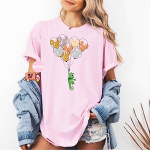 Może przedstawiać: R&oacute;żowy T-shirt z zielonym kameleonem trzymającym wiązkę kolorowych balon&oacute;w. Balony są w kształcie uszu Myszki Miki.