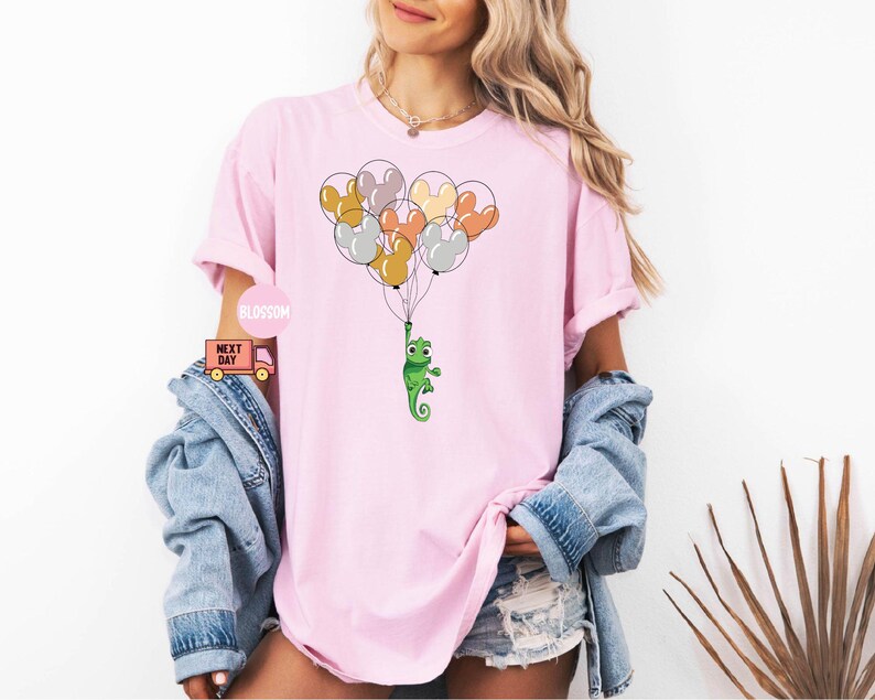 Może przedstawiać: R&oacute;żowy T-shirt z zielonym kameleonem trzymającym wiązkę kolorowych balon&oacute;w. Balony są w kształcie uszu Myszki Miki.