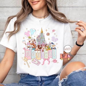 May include: White t-shirt featuring a colorful graphic of Disney characters, a castle, and the word "magical." The design includes Mickey Mouse, Minnie Mouse, Donald Duck, and other beloved characters. The shirt has a crew neck and short sleeves.