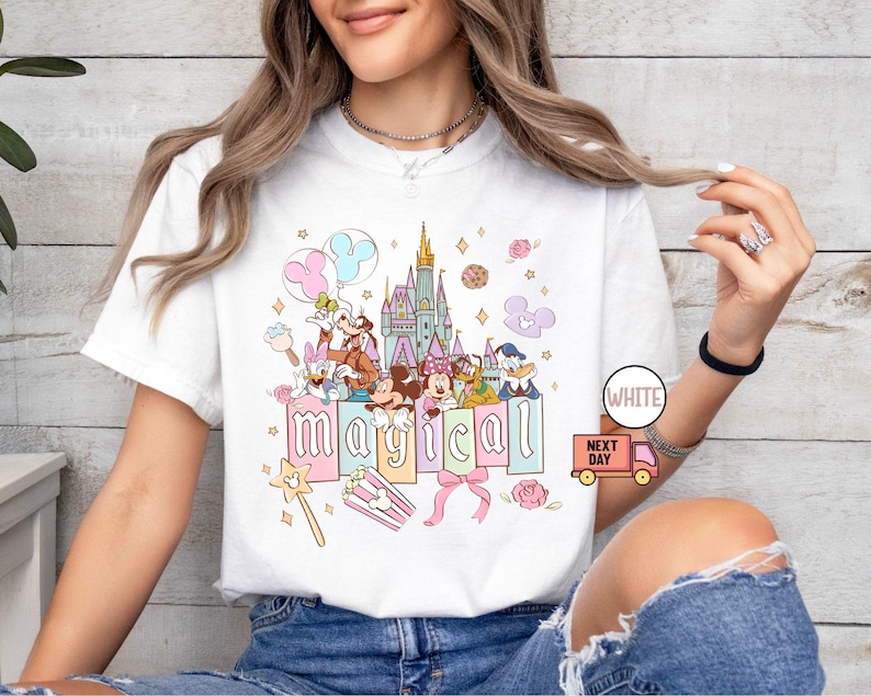 May include: White t-shirt featuring a colorful graphic of Disney characters, a castle, and the word "magical." The design includes Mickey Mouse, Minnie Mouse, Donald Duck, and other beloved characters. The shirt has a crew neck and short sleeves.
