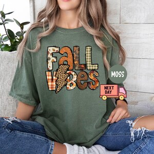 May include: Green t-shirt featuring the text "Fall Vibes" in a patchwork style with autumnal colours and patterns. The text is in a bold, blocky font.