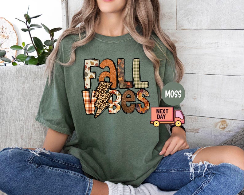 May include: Green t-shirt featuring the text "Fall Vibes" in a patchwork style with autumnal colours and patterns. The text is in a bold, blocky font.