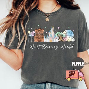 May include: Dark gray t-shirt featuring a colorful illustration of the Walt Disney World skyline. The design includes iconic landmarks like Cinderella Castle and Spaceship Earth, with the words "Walt Disney World" in a script font.