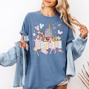 May include: Blue t-shirt with a colorful graphic featuring cartoon characters, a castle, and the word "magical." The design includes pastel colors and whimsical elements like a wand and popcorn.