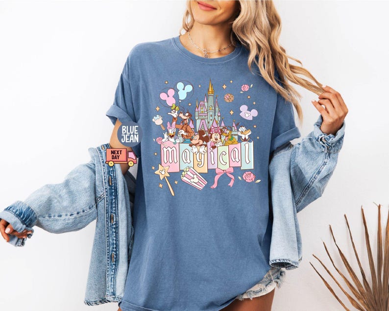 May include: Blue t-shirt with a colorful graphic featuring cartoon characters, a castle, and the word "magical." The design includes pastel colors and whimsical elements like a wand and popcorn.