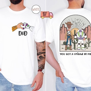 Pu&ograve; includere: Una t-shirt bianca con una grafica frontale di una mano umana e una mano robotica che formano un cuore sopra la scritta DAD. La grafica sul retro mostra due personaggi dei cartoni animati in un deserto con la scritta YOU GOT A FRIEND IN ME.