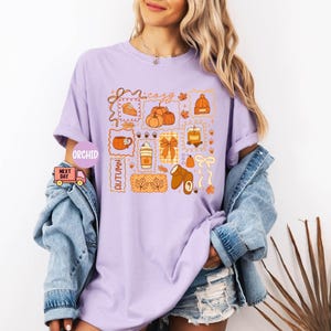 May include: A light purple t-shirt with a colorful autumn-themed graphic. The graphic includes pumpkins, a slice of pie, a coffee cup, and the word "cozy". The shirt is paired with a denim jacket and shorts.