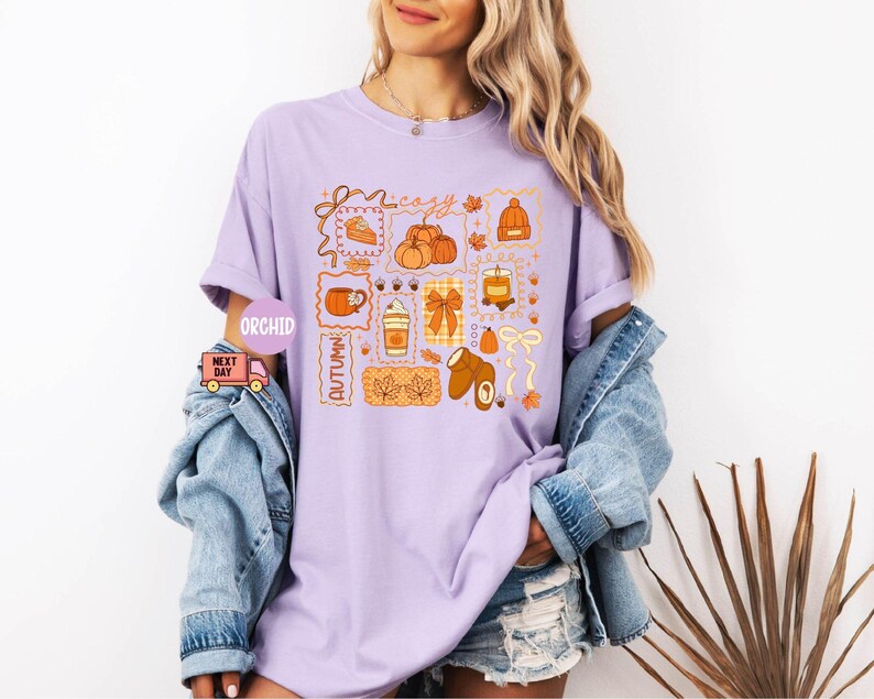 May include: A light purple t-shirt with a colorful autumn-themed graphic. The graphic includes pumpkins, a slice of pie, a coffee cup, and the word "cozy". The shirt is paired with a denim jacket and shorts.