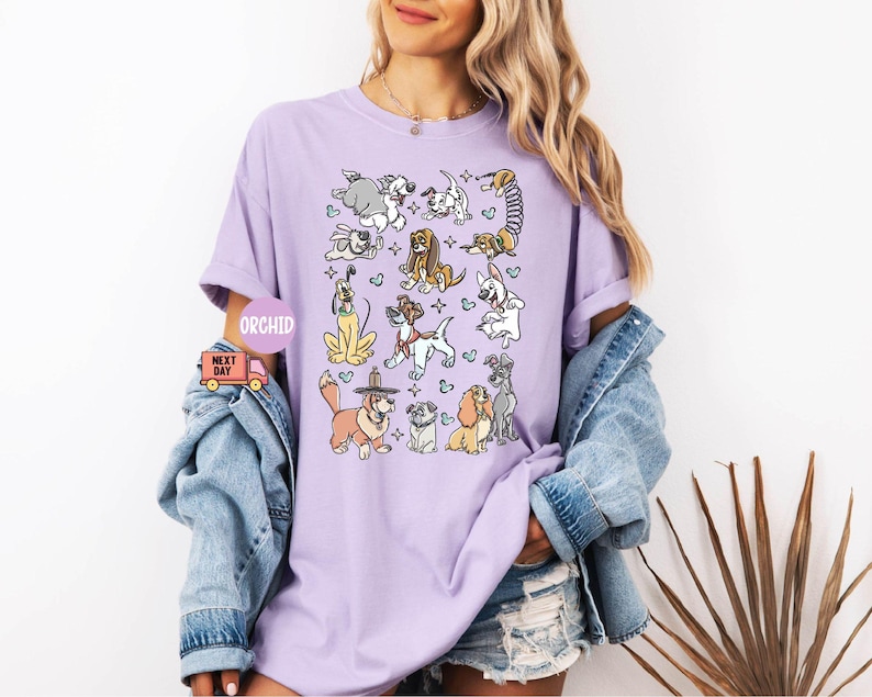May include: A light purple t-shirt featuring a colorful cartoon illustration of various dog breeds. The graphic includes dogs in different poses and styles, with hearts and stars. The word "ORCHID" is printed on the shirt.